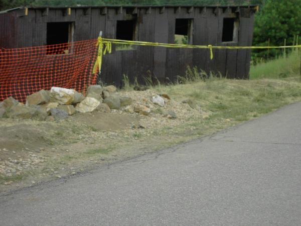 A rundown wooden structure with several open windows, surrounded by orange safety fencing and a pile of rocks. The scene is set against a grassy area next to a paved road. Betasso Preserve mountain bike trail.