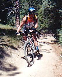 A cyclist riding a mountain bike along a dirt trail in a forested area, wearing a helmet and cycling gear. The scene shows greenery on either side of the path, indicating an outdoor setting. Betasso Preserve mountain bike trail.