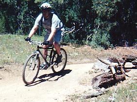 A person riding a mountain bike on a dirt trail surrounded by grass and trees, navigating around a log on the path. The cyclist is wearing a helmet and casual athletic clothing. Betasso Preserve mountain bike trail.