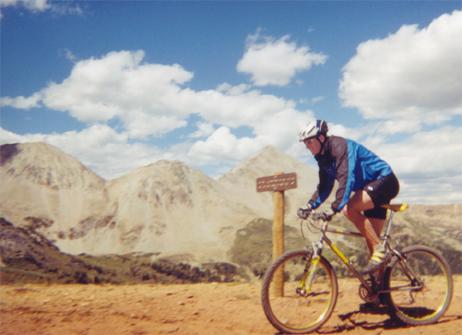 A mountain biker wearing a helmet and blue jacket rides along a dirt path with mountainous terrain in the background under a partly cloudy sky. A wooden signpost is visible beside the trail. Reno / Flag / Bear / Deadman Loop mountain bike trail.