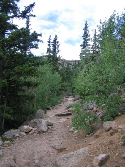 A dirt trail winding through a forested area, bordered by tall green trees and rocky terrain. The sky above is partially cloudy, suggesting a calm day in a natural setting. Barr Trail / Pikes Peak mountain bike trail.