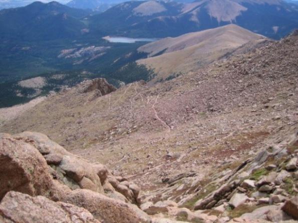 A panoramic view from a mountain slope, showcasing a rocky terrain with scattered boulders and a winding path leading downwards. In the distance, a lush green valley is visible, along with a lake nestled among the hills, under a clear blue sky and mountains. Barr Trail / Pikes Peak mountain bike trail.