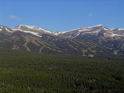 A panoramic view of a mountain range featuring snow-capped peaks and a dense forest below. The sky is clear, showcasing the natural beauty of the landscape, with various shades of green in the trees and rugged terrain. Wheeler National Recreation Trail mountain bike trail.