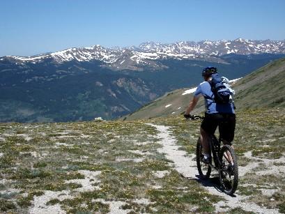A mountain biker riding along a narrow trail on a grassy hillside, with snow-capped peaks in the background under a clear blue sky. Wheeler National Recreation Trail mountain bike trail.