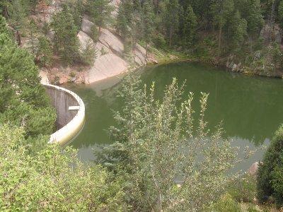 A serene landscape featuring a circular dam curving into a greenish lake, surrounded by lush trees and rocky hills in a natural setting. Winding Stairs Trail mountain bike trail.