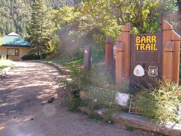 A well-maintained trailhead sign for Barr Trail, surrounded by greenery and wildflowers, with a nearby building featuring a blue roof. The path leads into a forested area, indicating access to hiking trails. Barr Trail / Pikes Peak mountain bike trail.