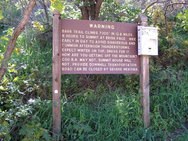 A wooden warning sign at the start of the Barr Trail, detailing important hiking information. The sign states that the trail climbs 7,300 feet over 12.6 miles and recommends an 8-hour hike to the summit. It advises hikers to begin early to avoid thunderstorms and to prepare for winter conditions at higher elevations. Additionally, it mentions that downhill transportation is not provided. The area is surrounded by greenery and trees. Barr Trail / Pikes Peak mountain bike trail.