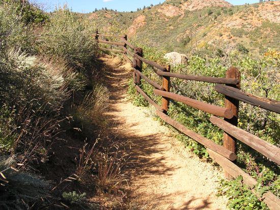 A dirt trail winds through a scenic landscape, bordered by a wooden fence. The path is surrounded by shrubs and grasses, with hills and rocky formations visible in the background under a clear blue sky. Barr Trail / Pikes Peak mountain bike trail.