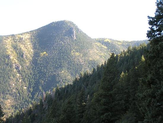 A scenic view of rugged mountains covered in dense evergreen forests, with a prominent peak rising in the background. The foreground features a lush treeline, while the mountains display hints of golden foliage, suggesting the transition of seasons. The sky is bright, illuminating the landscape. Barr Trail / Pikes Peak mountain bike trail.