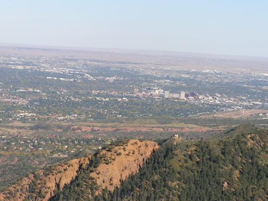 A panoramic view of a sprawling cityscape located in a valley, surrounded by distant mountains and hills. The foreground features rugged terrain with greenery, while the background showcases a mix of urban buildings and open land under a clear blue sky. Barr Trail / Pikes Peak mountain bike trail.