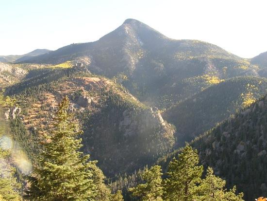 A sweeping mountain landscape featuring a prominent peak surrounded by lush green forests. The scene captures a valley with varied elevations, showcasing the natural beauty of the terrain under clear skies. Barr Trail / Pikes Peak mountain bike trail.