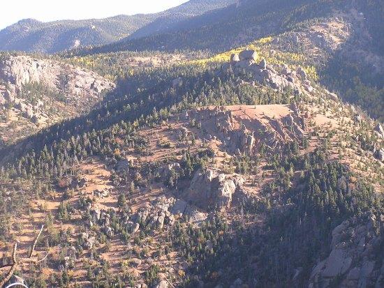A scenic view of rugged mountains covered with patches of trees and rocky outcrops, showcasing a landscape of varied terrain and natural beauty. The background features distant mountains, while the foreground highlights the texture of the rocks and the greenery of the forested areas. Bright sunlight illuminates parts of the landscape, creating a striking contrast between the natural elements. Barr Trail / Pikes Peak mountain bike trail.