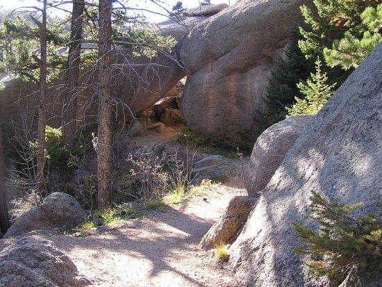 A narrow dirt path winding through a rocky landscape, flanked by trees and large granite boulders. Sunlight filters through the branches, illuminating the natural features of the area. Barr Trail / Pikes Peak mountain bike trail.