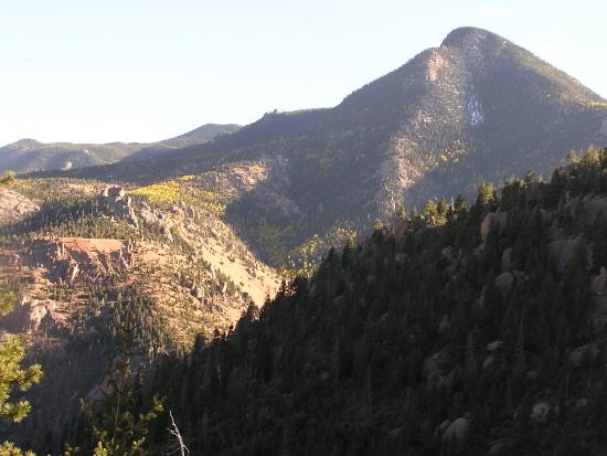 Mountain landscape featuring rolling hills and peaks, partially covered with trees, under a clear sky. The scene captures the natural beauty of mountainous terrain with varied elevations and textures. Barr Trail / Pikes Peak mountain bike trail.