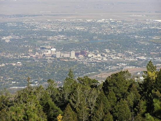 A panoramic view of a city surrounded by greenery, showcasing urban buildings and structures nestled among trees. The skyline reflects a mix of natural and urban landscapes, with distant hills and valleys visible in the background. Barr Trail / Pikes Peak mountain bike trail.