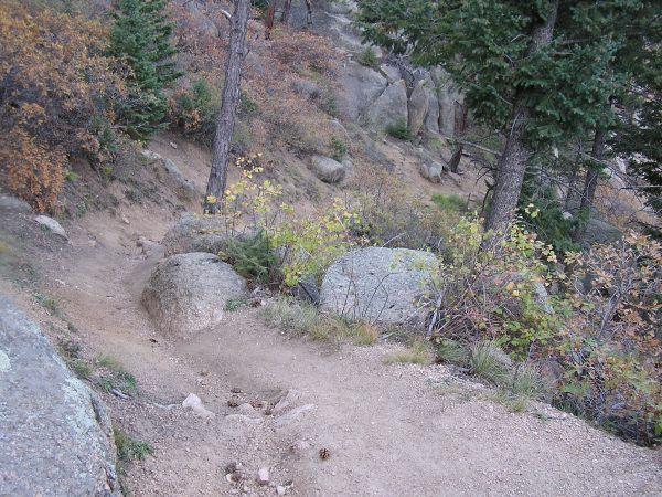 A winding dirt trail surrounded by rocks, shrubs, and trees, depicting a serene natural landscape. The scene includes various shades of green and autumn colors in the foliage, suggesting a forested area. Barr Trail / Pikes Peak mountain bike trail.