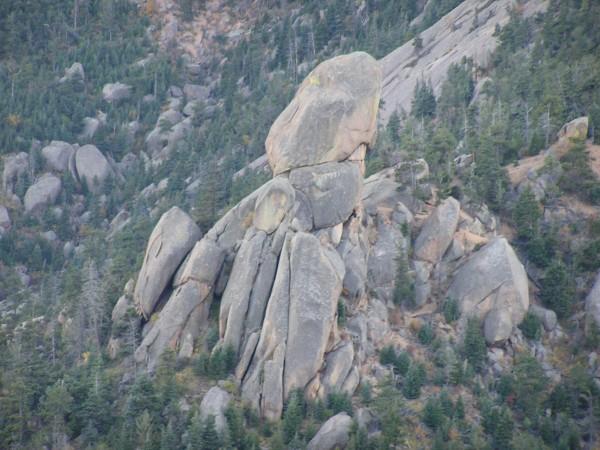 A cluster of large, rugged boulders is situated amidst a backdrop of dense evergreen trees on a rocky hillside. The boulders vary in size and shape, with one prominently perched atop a larger formation, creating a striking natural sculpture. The scene reflects the raw beauty of mountainous terrain. Barr Trail / Pikes Peak mountain bike trail.