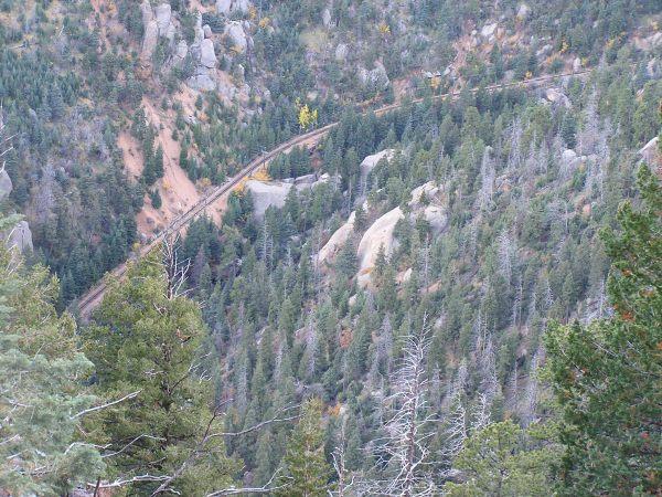 Aerial view of a winding railway track meandering through a dense forest of evergreen trees, with rocky formations visible in the background. The scene conveys a sense of natural beauty and rugged terrain. Barr Trail / Pikes Peak mountain bike trail.