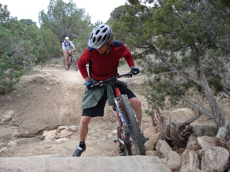 A mountain biker navigates a rocky trail, adjusting their bicycle while wearing a helmet and a red long-sleeve shirt. Another cyclist is seen in the background, riding along the path through a wooded area. Prime Cut mountain bike trail.