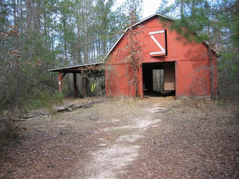An old, weathered red barn is situated in a clearing surrounded by tall trees. The path leading to the barn is partially overgrown with leaves and branches. The barn's front doors are slightly ajar, revealing a dark interior, and a small roof extension is visible on one side. Sunlight filters through the trees, casting dappled light on the scene. Arrowhead Park mountain bike trail.