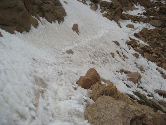 Snow and rocky terrain on a mountainside, with patches of snow and scattered boulders. The snow appears uneven and textured, contrasting with the surrounding rocky surfaces. Barr Trail / Pikes Peak mountain bike trail.