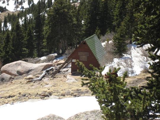 A small, rustic cabin with a green roof sits among rocky terrain and patches of snow, surrounded by evergreen trees. The scene captures a serene, mountainous landscape, indicating a remote and tranquil environment. Barr Trail / Pikes Peak mountain bike trail.