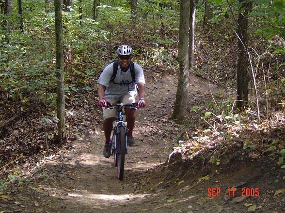 A person riding a mountain bike on a dirt path through a forested area, surrounded by trees and foliage. The cyclist is wearing a helmet and sunglasses, and is dressed in a t-shirt and shorts. The image captures the outdoor environment with dirt trail winding through the woods. The date at the bottom indicates September 17, 2005. Fort Custer Recreation Area mountain bike trail.