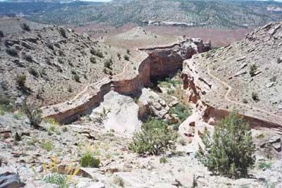 A barren landscape featuring a deep canyon winding through rocky terrain, surrounded by sparse vegetation and distant hills under a clear blue sky. Kokopelli Trail mountain bike trail.