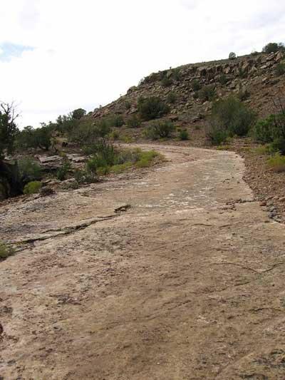 A rocky trail winding through a sparse, rugged landscape, with a hill in the background and scattered vegetation along the sides. The sky is partly cloudy, creating a natural and serene outdoor setting. Kokopelli Trail mountain bike trail.