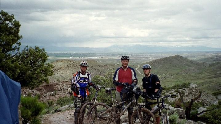 Three mountain bikers pose on a rocky outcrop overlooking a scenic valley. They are wearing helmets and biking gear, with their mountain bikes beside them. The landscape features green hills and a cloudy sky, indicating an outdoor adventure setting. Kokopelli Trail mountain bike trail.