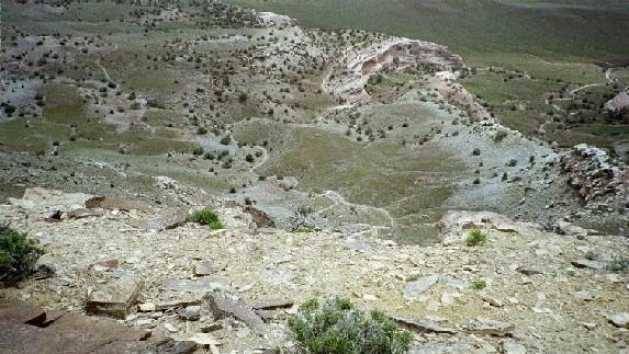 A panoramic view of a rocky landscape showcasing a valley with sparse vegetation, rolling hills, and a winding path. The foreground features large stones and small plants, while the background reveals a mixture of green and rocky terrain. The scene conveys a sense of natural beauty and rugged terrain. Kokopelli Trail mountain bike trail.