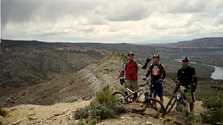Three mountain bikers pose at the edge of a rocky cliff, overlooking a scenic valley below. They wear safety gear, including helmets and pads, and hold their bikes. The landscape features rolling hills and a river visible in the distance under a cloudy sky. Kokopelli Trail mountain bike trail.