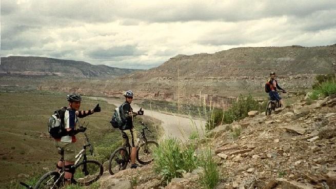 Three mountain bikers on a rocky trail overlook a scenic landscape with hills and a river below. Two of the bikers give a thumbs-up gesture while standing beside their bikes, and one biker sits on a bike further down the trail. The sky is partly cloudy, creating a dramatic backdrop for their outdoor adventure. Kokopelli Trail mountain bike trail.