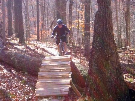 A mountain biker riding over a wooden bridge in a wooded area during autumn. Trees are bare, and fallen leaves cover the ground. The sunlight filters through the branches, creating a warm glow on the scene. Brown County Park mountain bike trail.