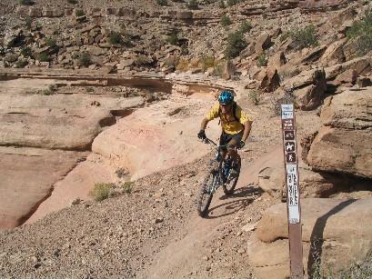 A mountain biker wearing a yellow shirt and blue helmet navigating a rocky trail. In the background, layered rock formations and sparse vegetation are visible, along with a trail marker sign indicating the path. Kokopelli Trail mountain bike trail.