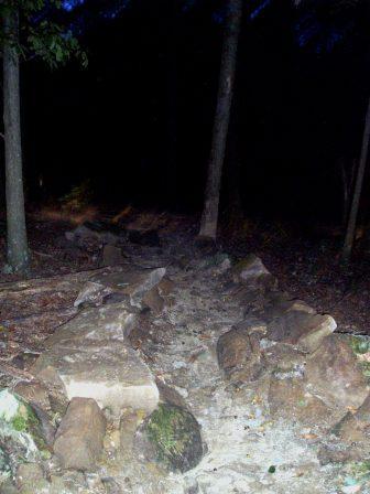 A dimly lit pathway in a wooded area, featuring scattered rocks and uneven terrain. Tall trees surround the path, creating a natural, rustic setting at night. Brown County Park mountain bike trail.