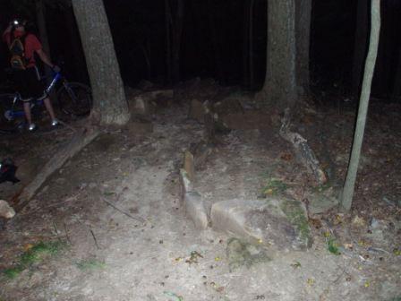 A dark forested area at night with visible rocks and a dirt path. A partially obscured person wearing a red shirt is standing next to a bicycle, surrounded by trees. Brown County Park mountain bike trail.