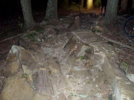 Rocky trail in a wooded area at night, showing a path made of large stones and dirt. Trees are visible on either side, with a faint light in the background suggesting human presence. Brown County Park mountain bike trail.