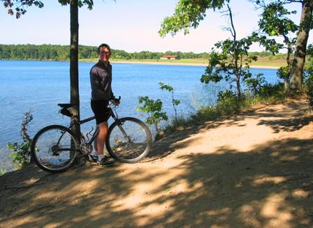 A person standing next to a mountain bike on a sandy path by a lake, surrounded by trees and greenery. The sun is shining, and there is a peaceful scene of water and distant hills in the background. Fort Custer Recreation Area mountain bike trail.