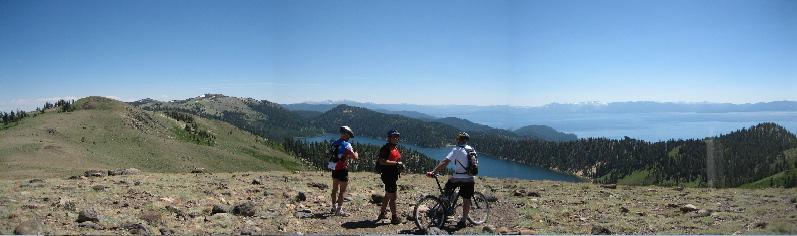 Three cyclists standing on a rocky hillside overlooking a scenic lake surrounded by mountains under a clear blue sky. The landscape features lush greenery and distant peaks, creating a beautiful backdrop for outdoor activities. Tahoe Rim Trail:  Kingsbury South To Armstrong Pass mountain bike trail.