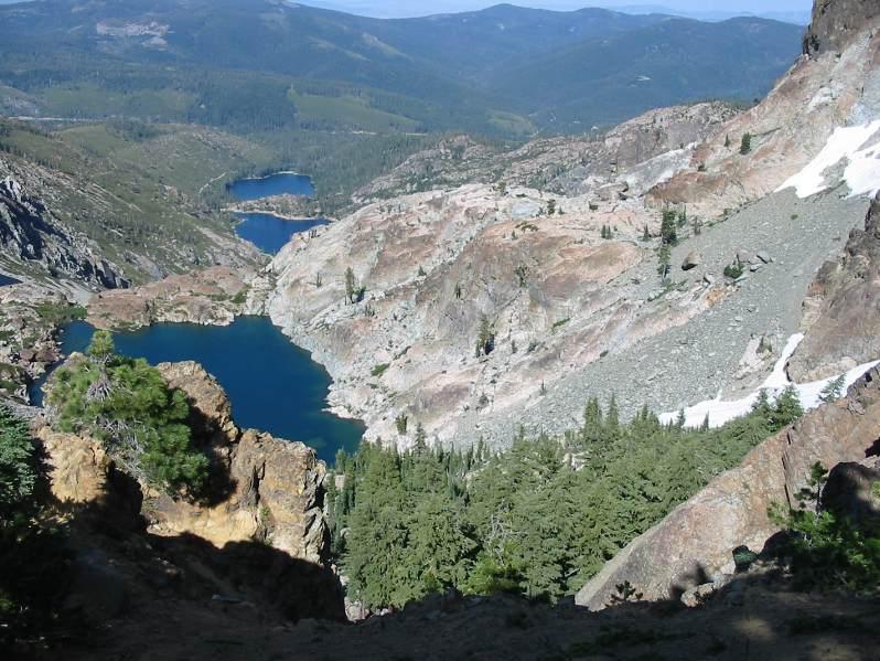 A scenic view of rugged mountainous terrain featuring two lakes nestled between rocky slopes. The foreground includes pine trees and rocky outcrops, with slopes displaying a mix of vegetation and bare rock. In the background, additional lush green mountains can be seen under a clear blue sky. Tahoe Rim Trail:  Kingsbury South To Armstrong Pass mountain bike trail.