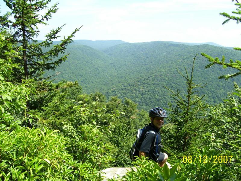 A person wearing a bike helmet and a backpack is sitting on a rock near the edge of a lush green forest, overlooking a scenic valley filled with rolling mountains. The scene is bright and sunny, showcasing vibrant foliage and a clear blue sky in the background. Gauley Headwaters mountain bike trail.
