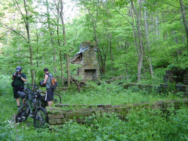 Two mountain bikers stand near their bikes in a lush, green forest. In the background, there is a partially collapsed stone structure, likely remnants of an old building, surrounded by dense vegetation and trees. The scene conveys a sense of exploration and the passage of time in nature. Brown County Park mountain bike trail.