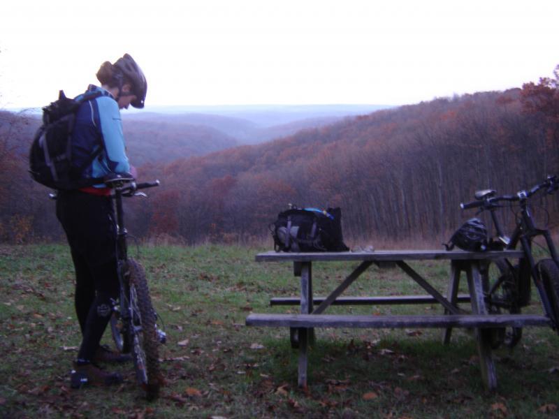 A person in cycling gear stands beside a mountain bike, checking their equipment on a scenic overlook. In the background, rolling hills covered with autumn foliage stretch into the distance. A picnic table and another bike are visible nearby, set against a tranquil landscape. Brown County Park mountain bike trail.