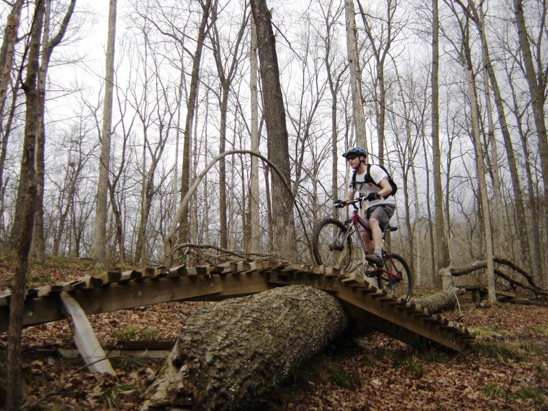 A mountain biker navigating a wooden ramp over a fallen log in a forested area with bare trees, showcasing an adventurous outdoor scene. Brown County Park mountain bike trail.