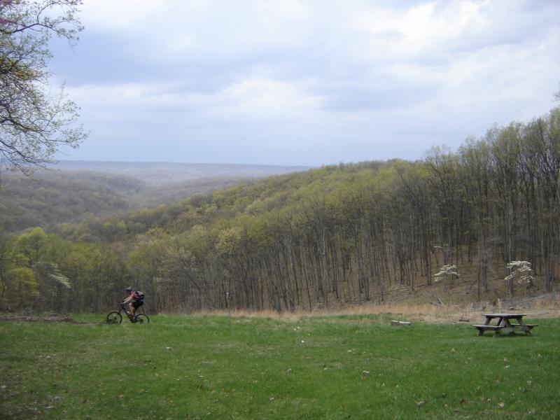 A mountain biker rides along a grassy area overlooking a hilly landscape with dense trees and a cloudy sky in the background. A picnic table is visible in the foreground. Brown County Park mountain bike trail.
