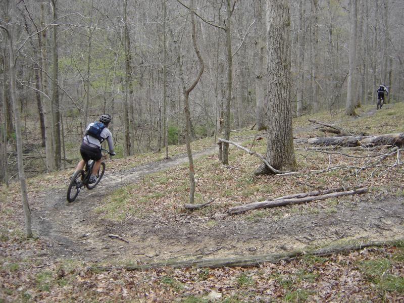 A mountain biker rides along a narrow dirt trail in a wooded area, surrounded by bare trees and scattered leaves on the ground. Another cyclist can be seen in the background on a separate path. Brown County Park mountain bike trail.