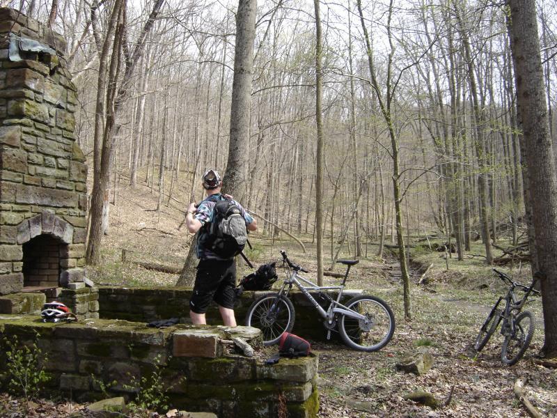 A person wearing a backpack stands on a stone structure in a wooded area, looking into the distance. Two bicycles are parked beside the stone structure, and the landscape features bare trees and a mix of rocky and grassy terrain. Brown County Park mountain bike trail.