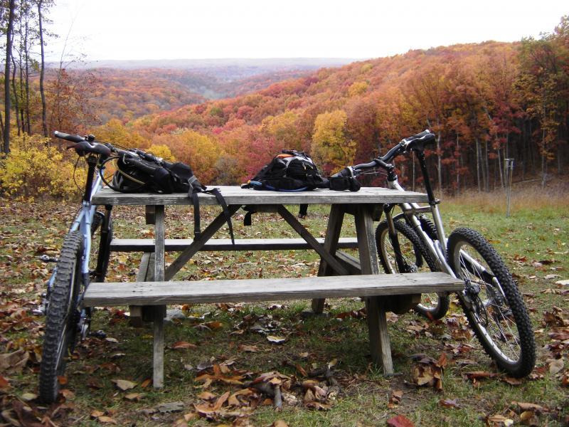 Two mountain bikes lean against a wooden picnic table, with backpacks placed on the table. In the background, a scenic view of rolling hills showcases vibrant fall foliage in shades of orange and red, surrounded by a clear sky. Fallen leaves cover the ground, adding to the autumnal atmosphere. Brown County Park mountain bike trail.