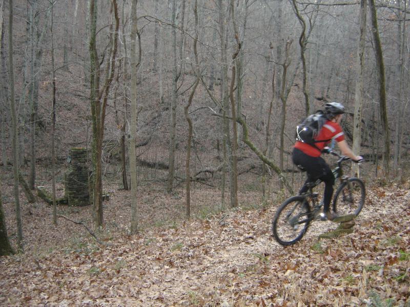 A mountain biker rides along a dirt path in a wooded area during autumn. The landscape features bare trees and fallen leaves, with a stone structure partially visible in the background. The cyclist is wearing a red shirt and a helmet, and is positioned on a slope amidst the serene natural setting. Brown County Park mountain bike trail.